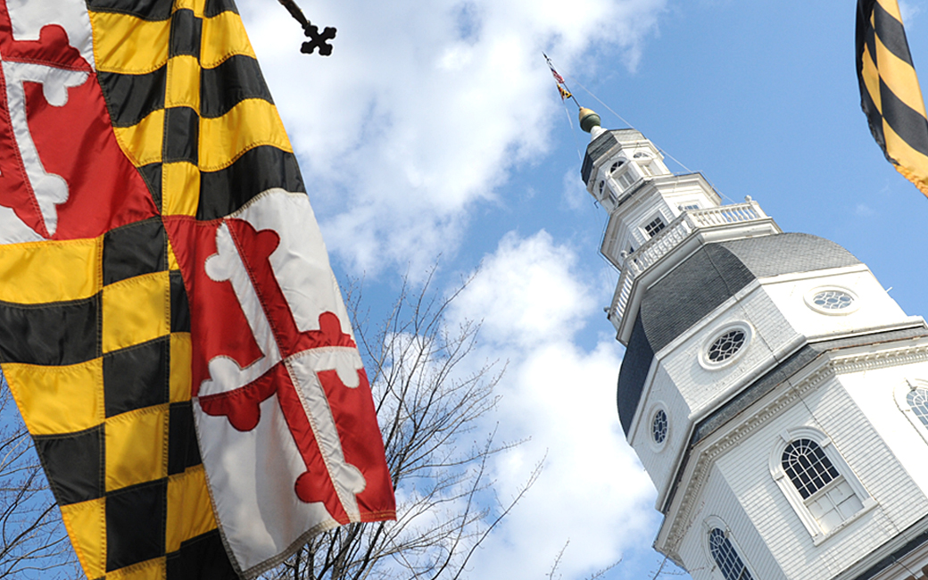 maryland state flag flying next to cupola of maryland state house with blue sky