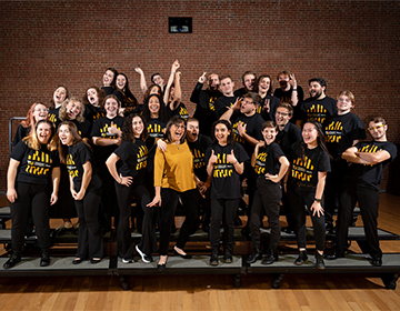 The choir smiles and makes silly faces at the camera from the Harold J Kaplan Concert Hall stage, with Professor Saez smiling in the middle