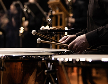 A drummer in black plays steady notes on large, shiny drums