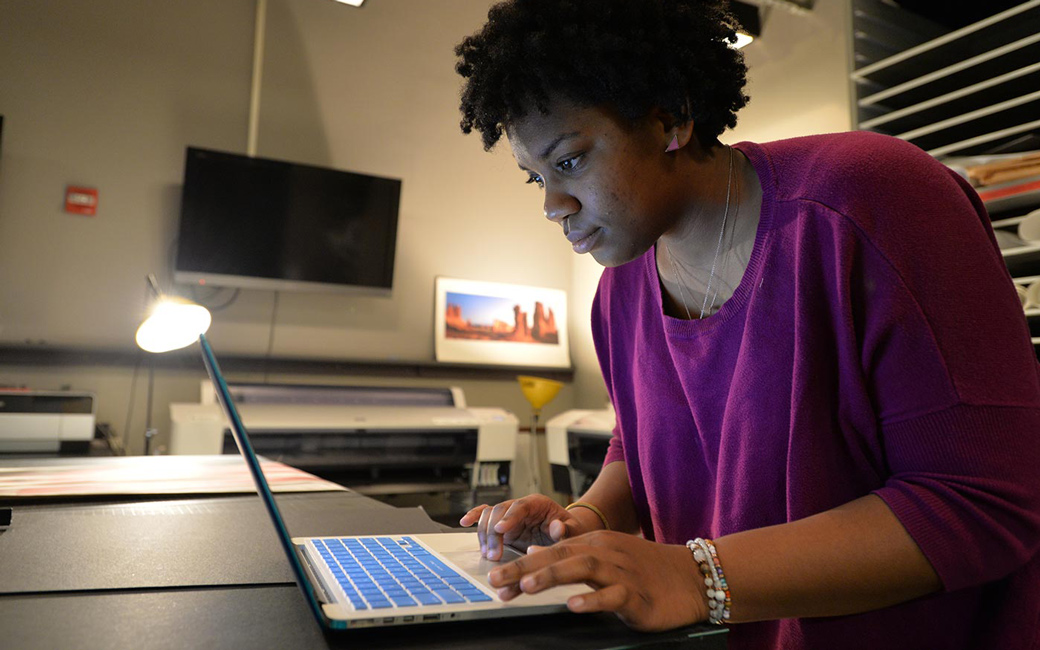 An MFA student navigates on her laptop, with artwork hanging in the background