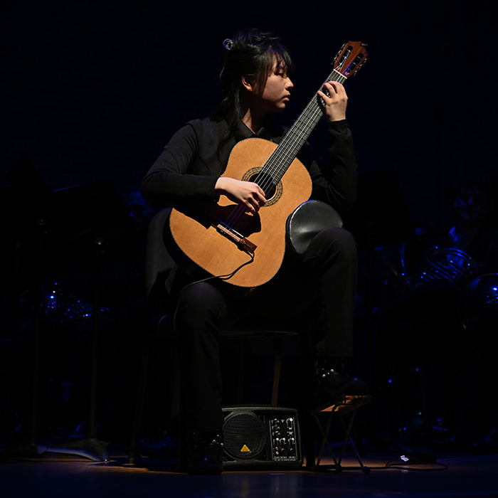 A musician in dark clothing and against a dark background plays a guitar -- she and the guitar are lit in stark contrast to everything else around her