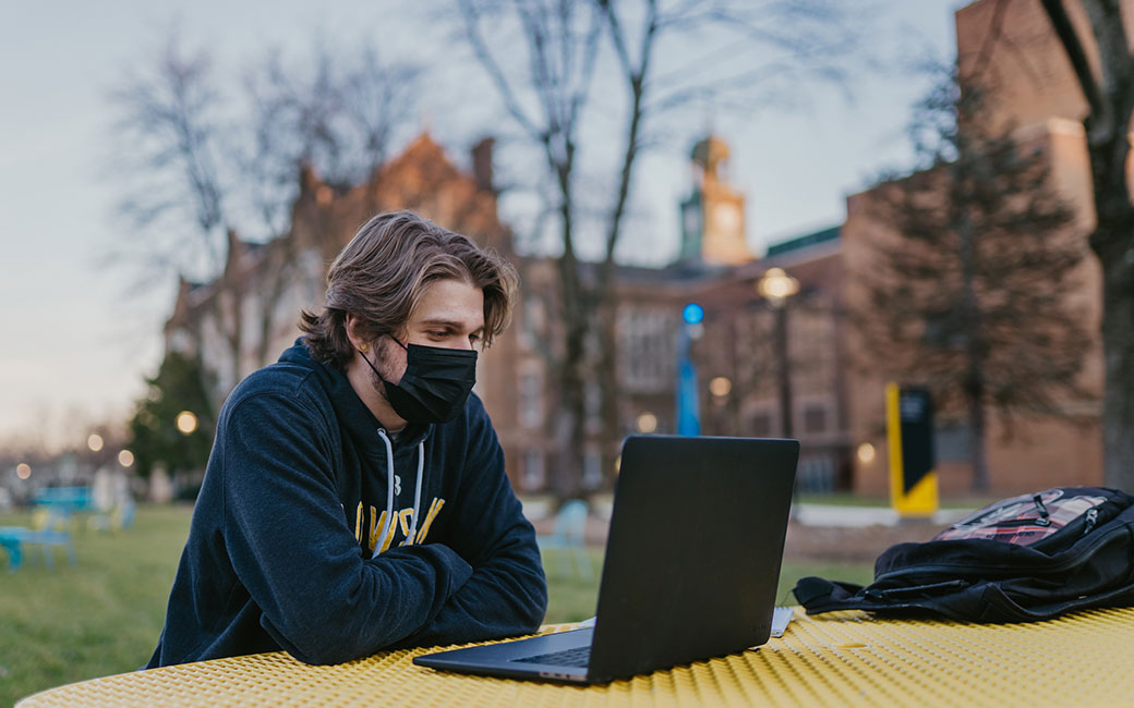 Masked student studying with laptop outside