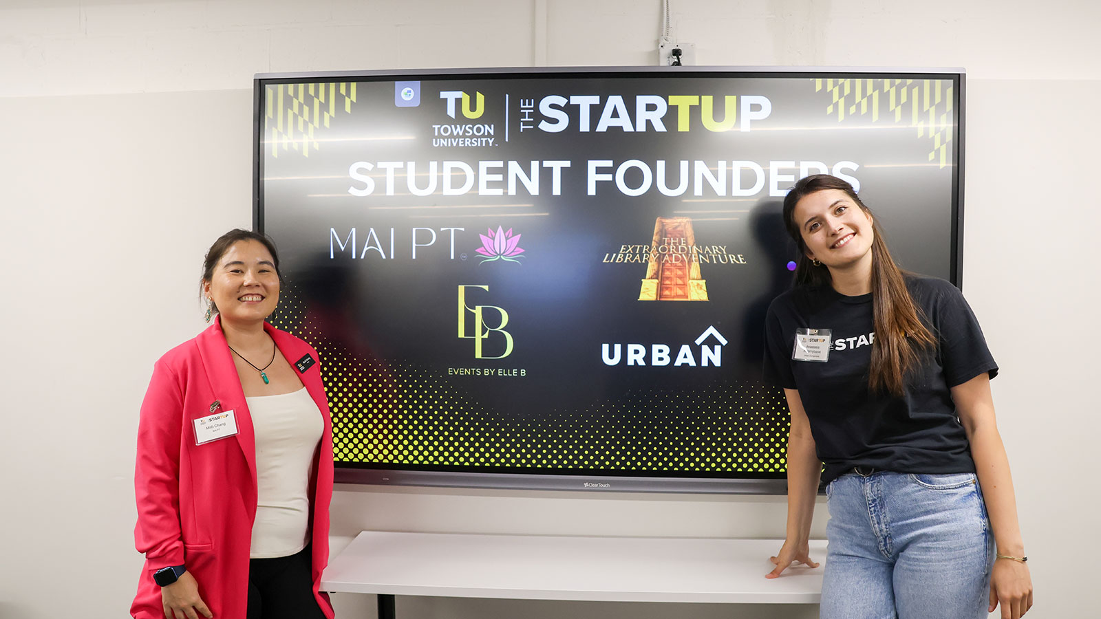 Two students in front of board that shows the logos for each student founder at the Accelerator