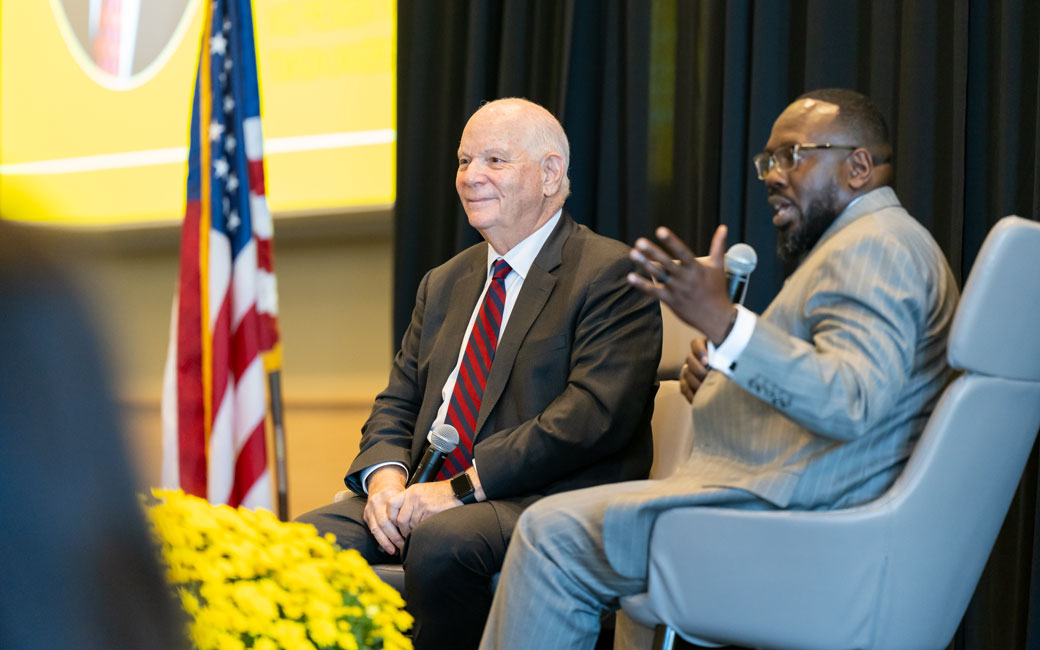 Two men sit in front of audience and facilitate a discussion.