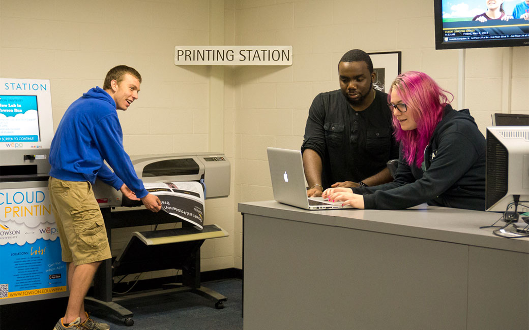 a student prints a poster while staff help with a laptop problem nearby